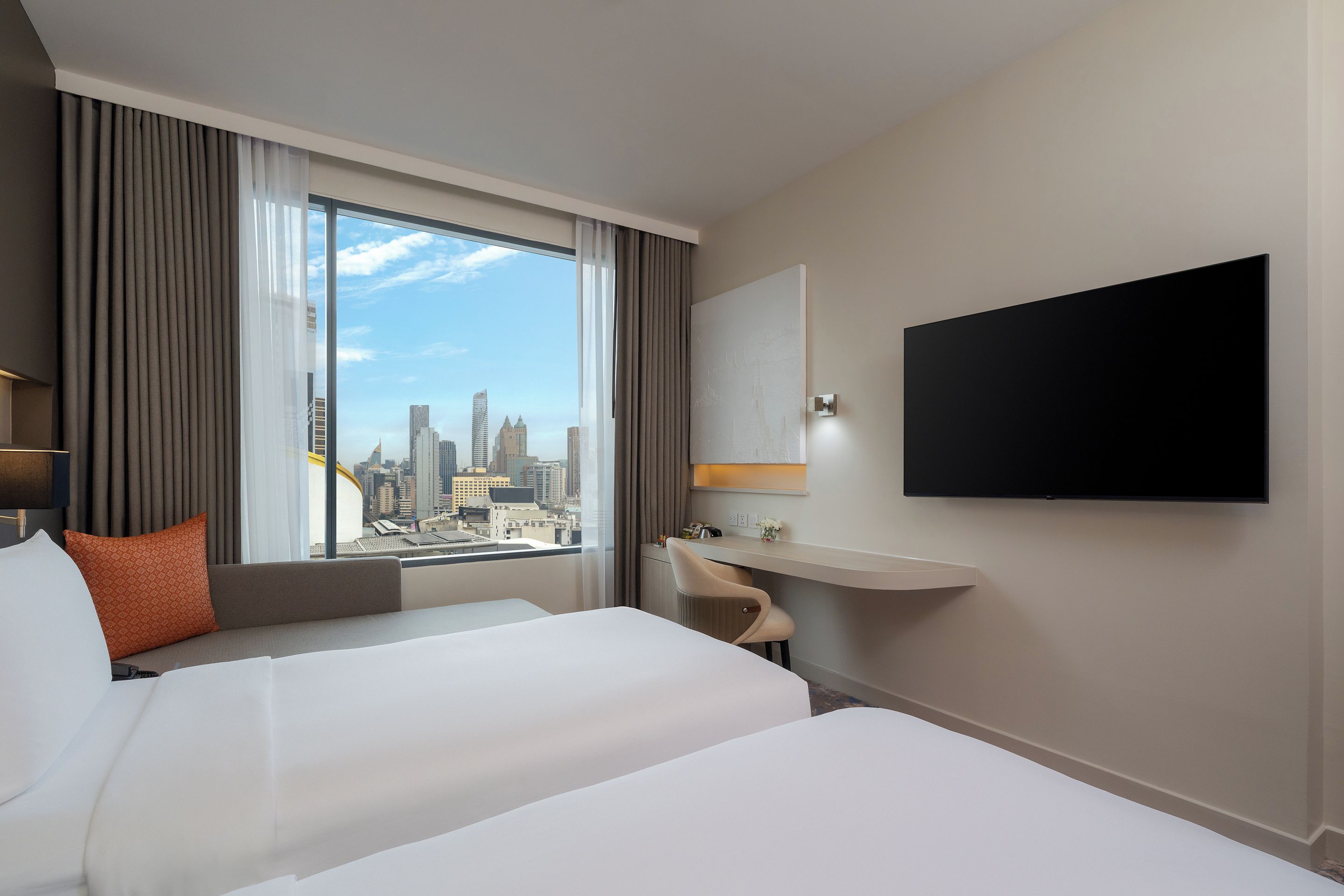 A hotel room with a bed, a desk, a chair, a TV, and a view of a cityscape.