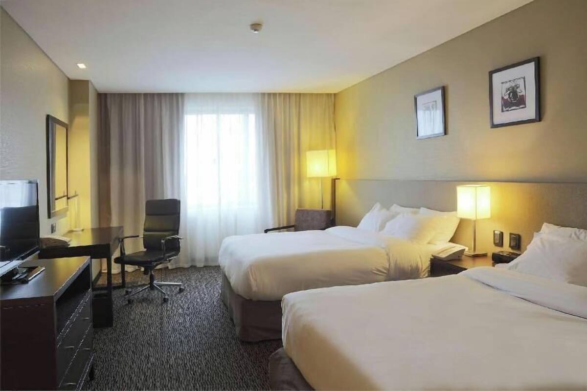 A hotel room with two beds, a desk with a chair, a television, and a large window with curtains.