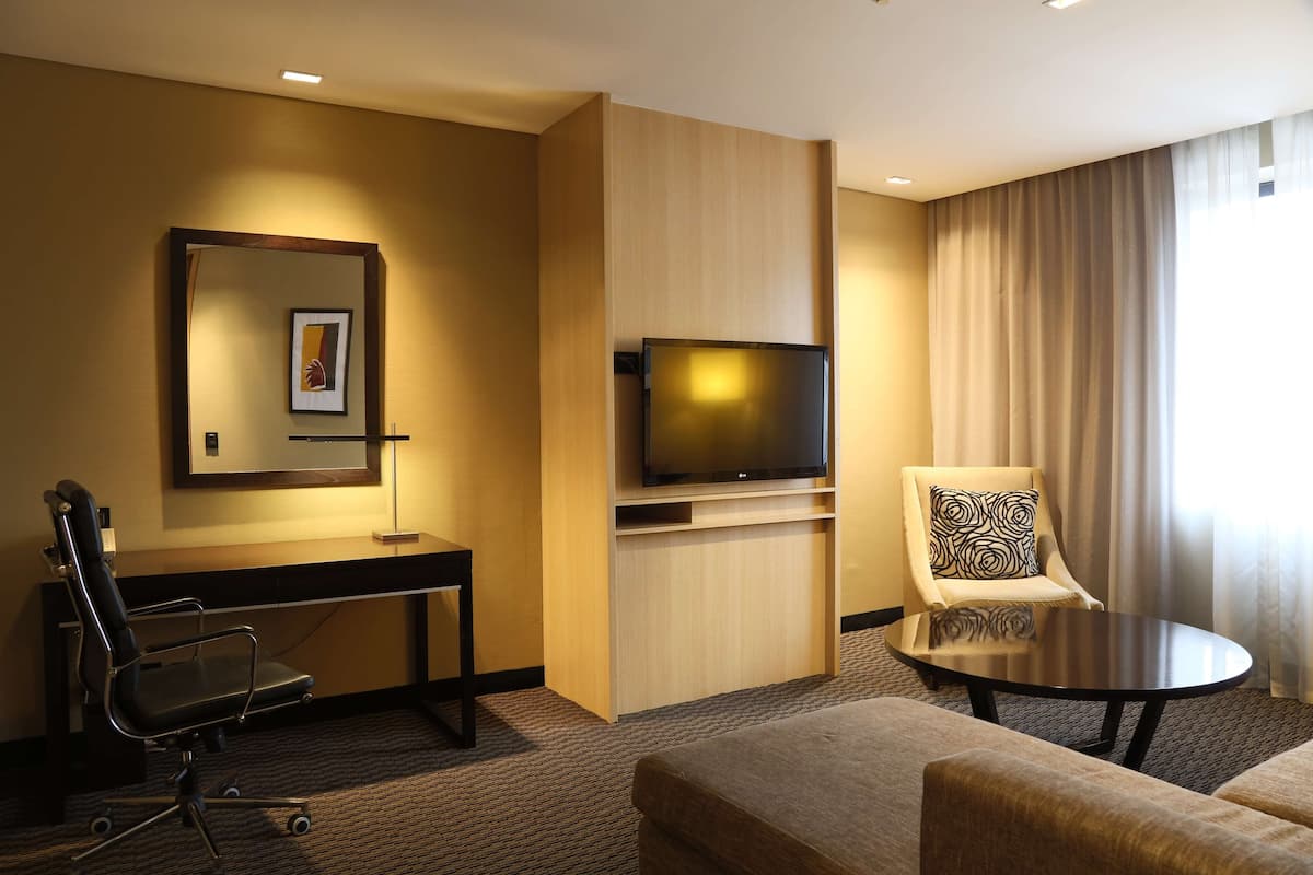 A hotel room with a desk, chair, TV, and a sofa.