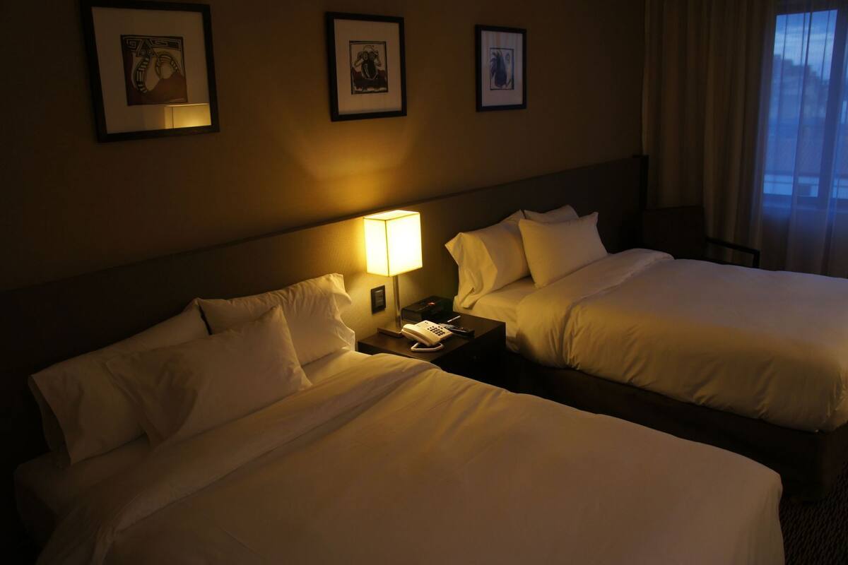 A hotel room with two beds, a nightstand with a lamp, and framed artwork on the wall.