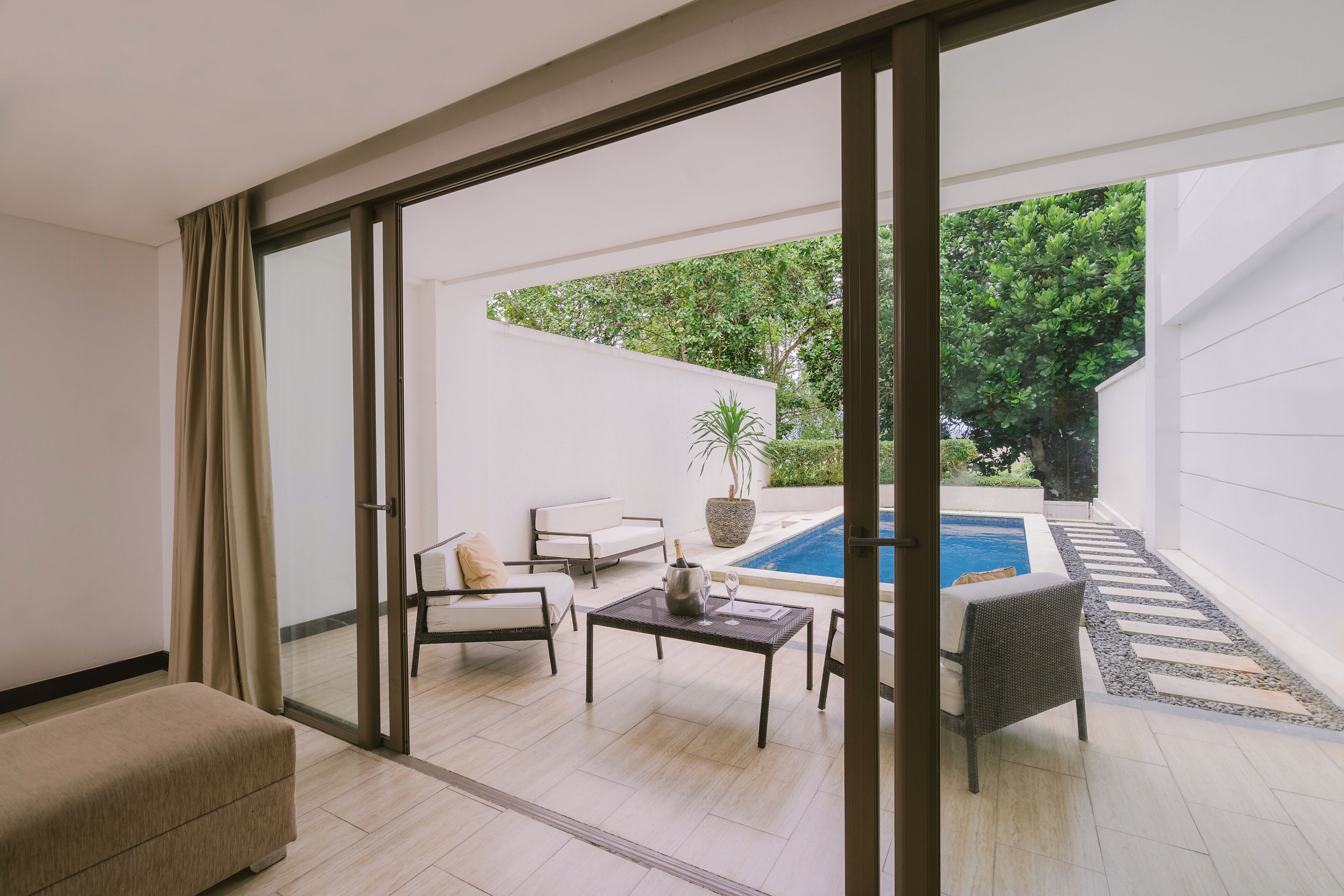 Un salon moderne doté d’une porte vitrée donnant sur une piscine, d’un canapé, d’une table basse et d’un fauteuil en osier.