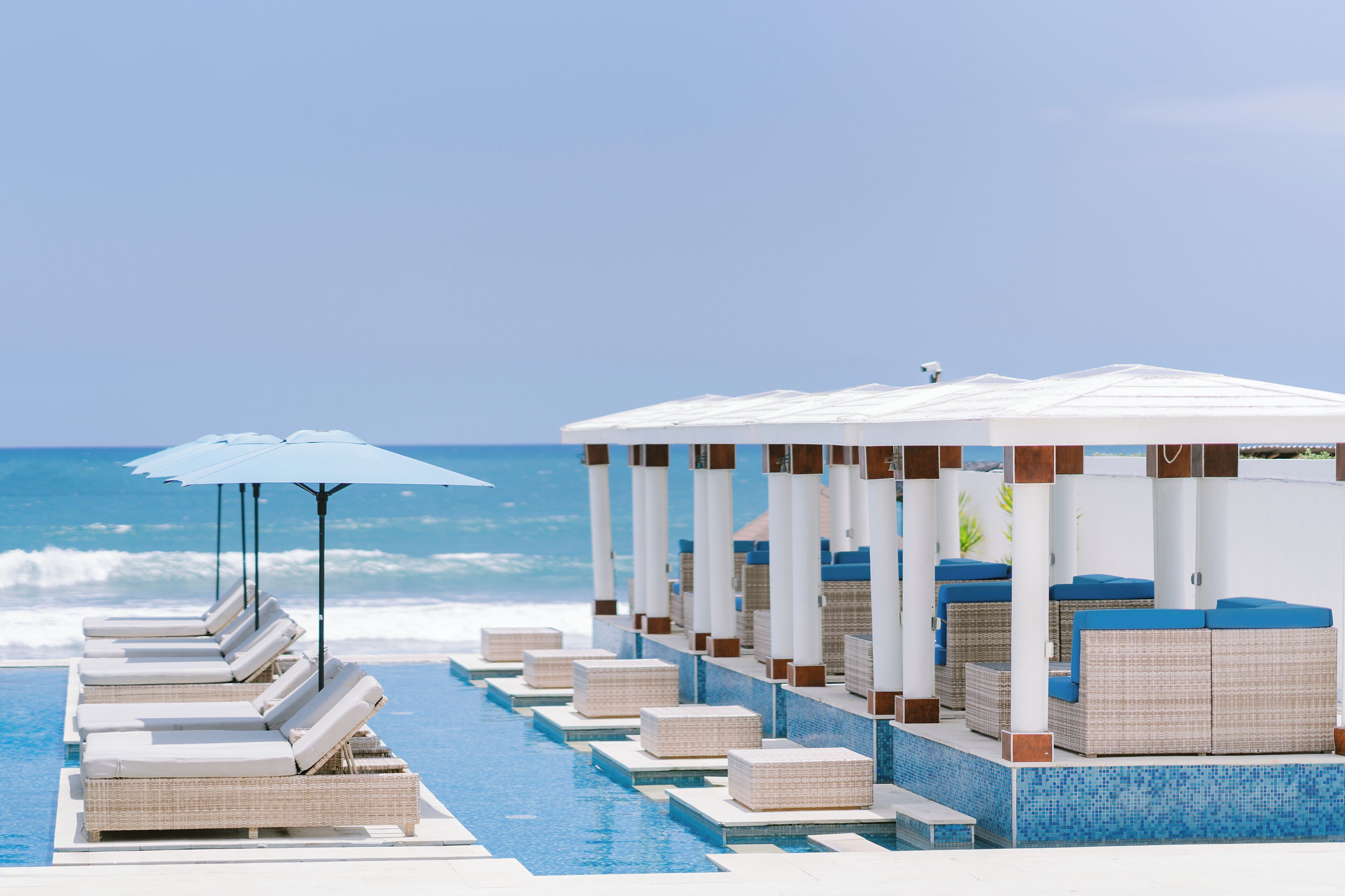 Un área junto a la piscina con sillones de descanso y sombrillas, un espacio de estar cubierto y vista al océano.