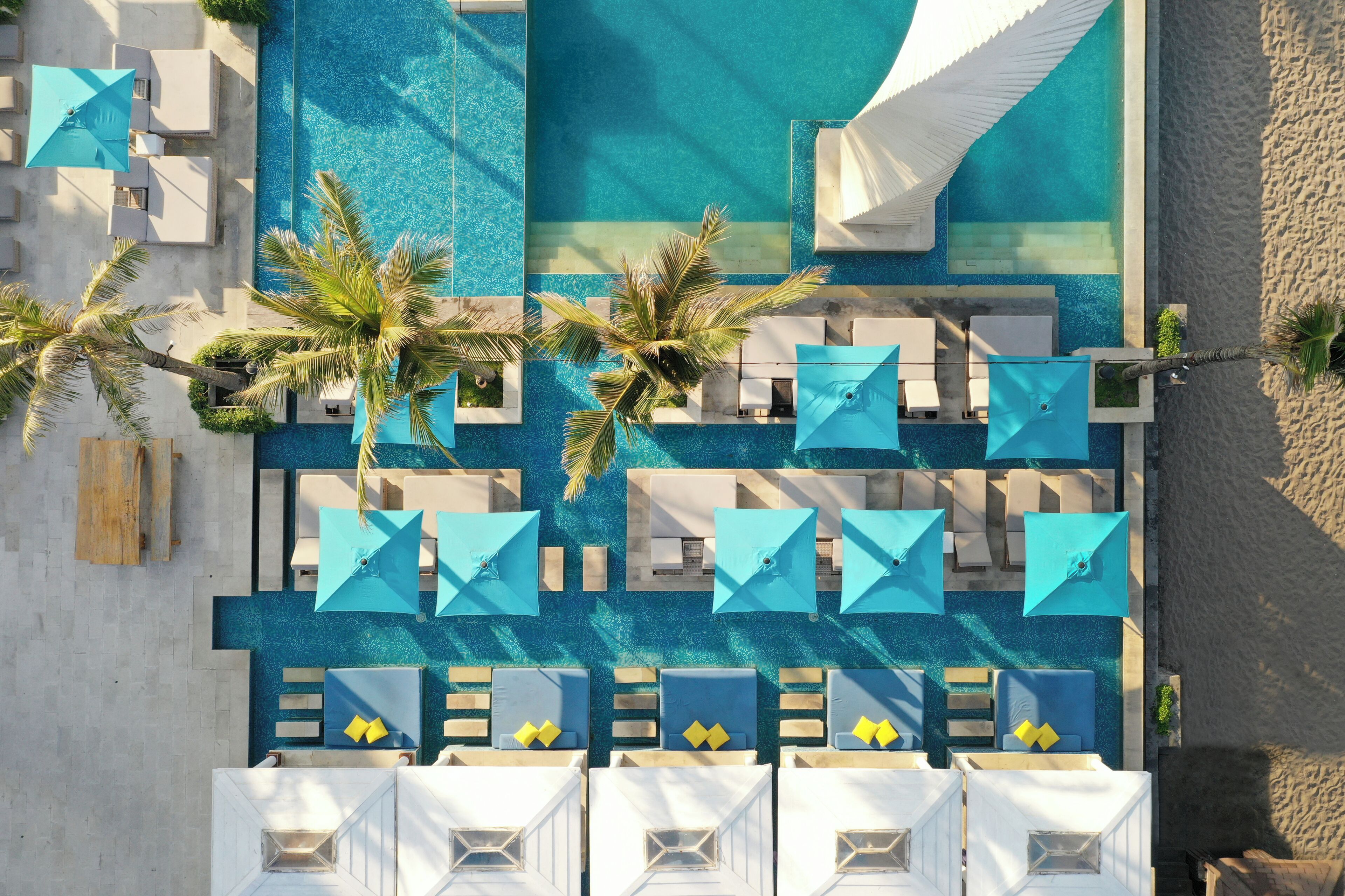Vue aérienne d’un complexe hôtelier avec une piscine, des chaises longues, des parasols et des palmiers.