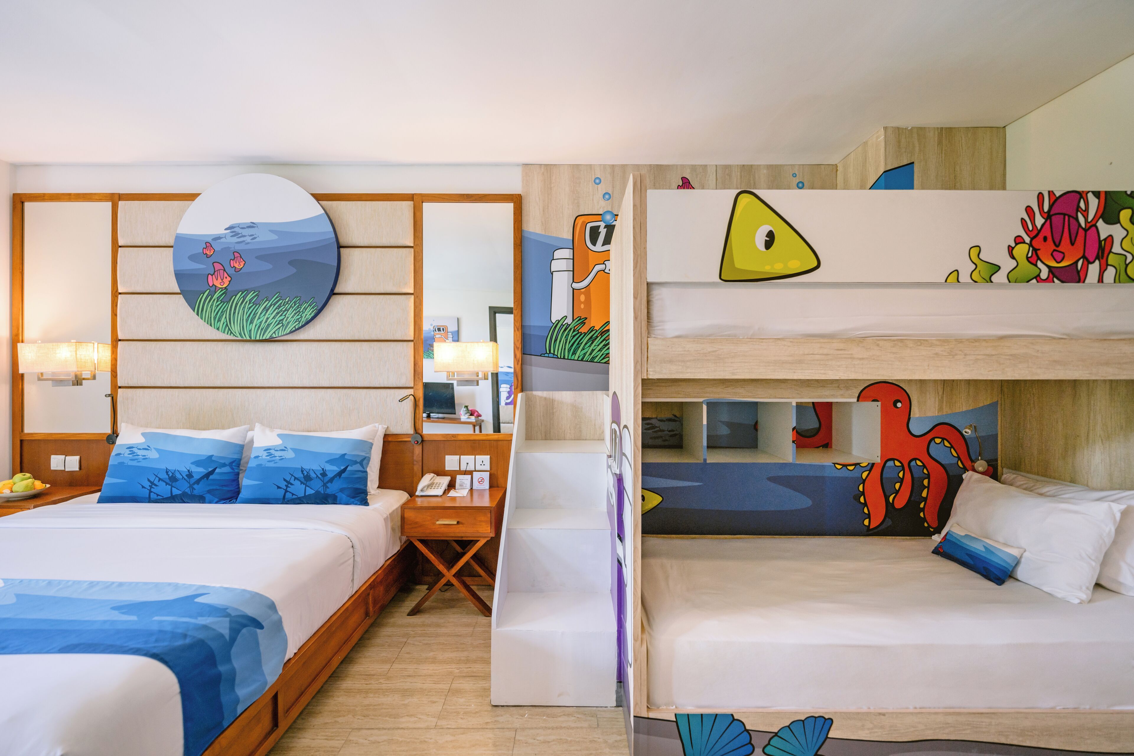 Quarto de hotel com duas camas, uma escrivaninha, uma cadeira, uma luminária e obras de arte nas paredes.