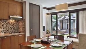 A modern dining area with a wooden table set for four, a kitchen with a tiled backsplash, and a view of a private outdoor seating area. - Seminyak Townhouse Apartment (Legian)
