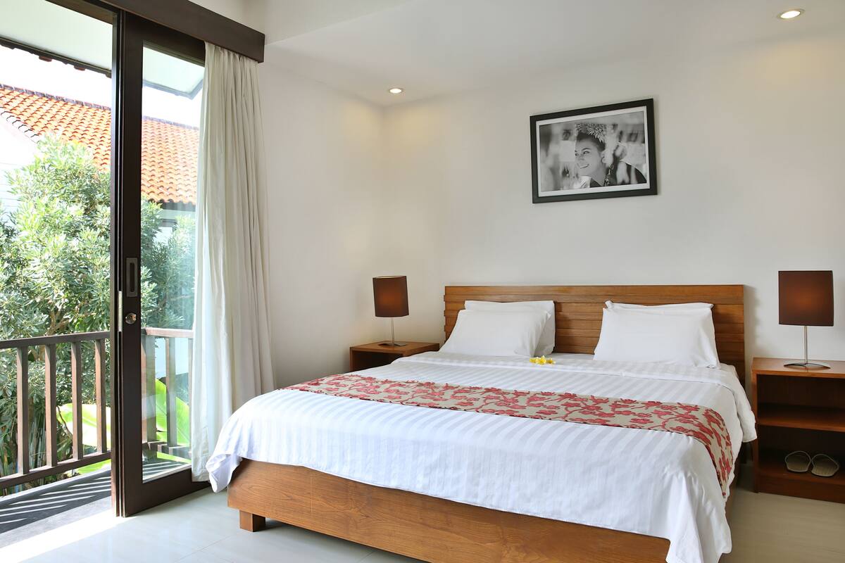 A bedroom with a bed, nightstands, a picture on the wall, and a balcony with a view of greenery.