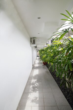 A corridor with tiled flooring, white walls, and potted plants along one side.