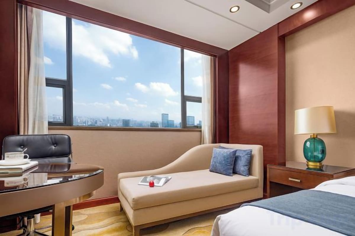 A hotel room with a large window, a sofa, a bed, a desk, and a lamp.