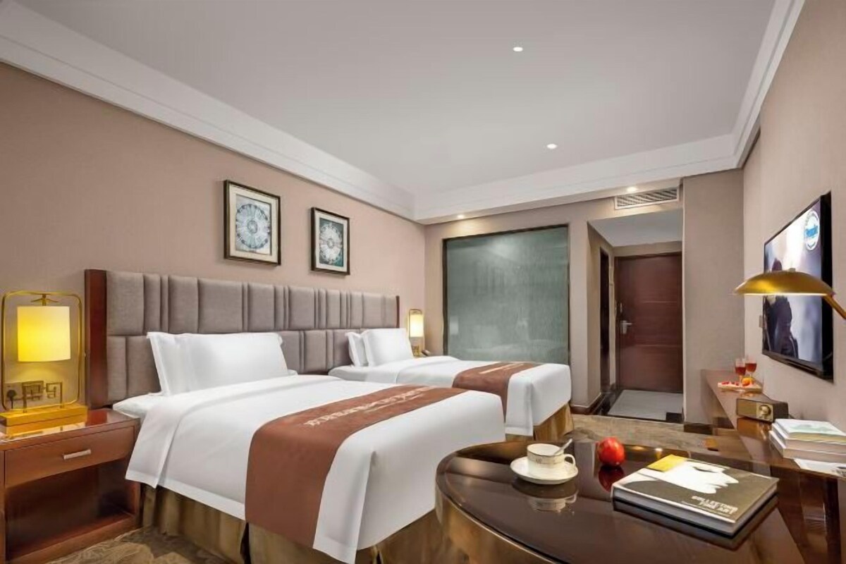 A hotel room with two beds, a desk, a chair, a lamp, and a television.