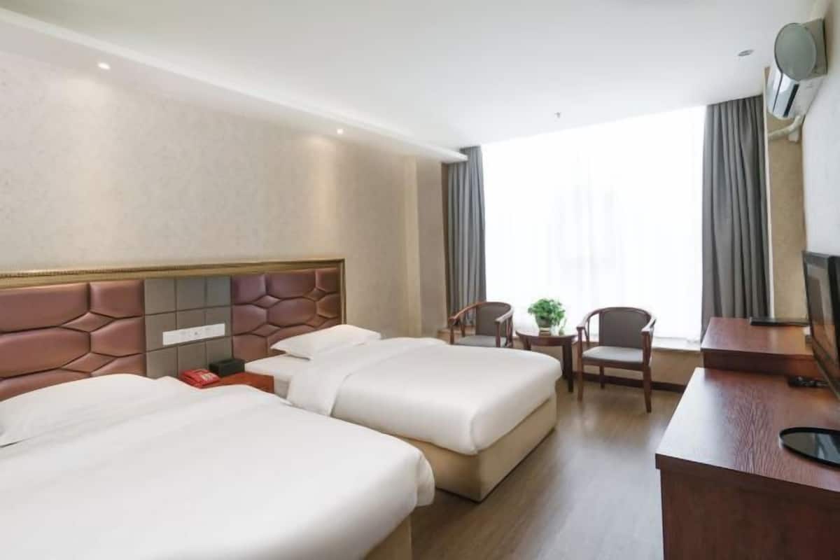 Ein Hotelzimmer mit zwei Betten, einem Schreibtisch, einem Stuhl, einem kleinen Tisch mit Pflanze und einem großen Fenster mit Vorhängen.