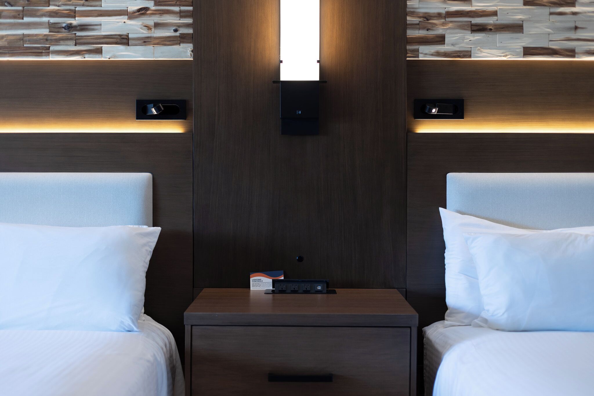 A hotel room with two beds, a nightstand, and a wall-mounted light fixture.