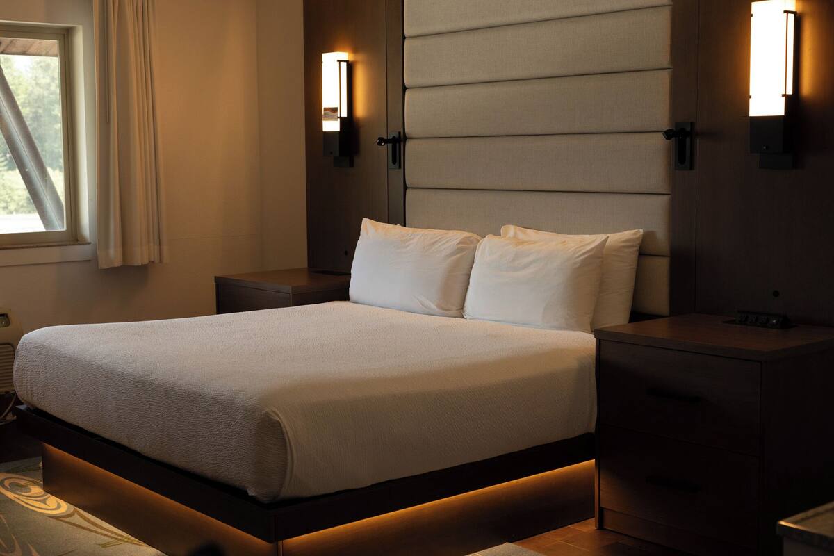 A neatly made bed with a white comforter and pillows, flanked by two bedside tables with lamps, in a modern room with a large window and a wall-mounted light fixture.