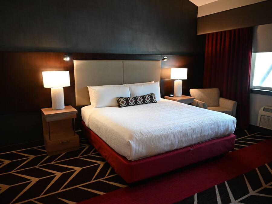 Habitación de hotel con una cama grande, dos lámparas de noche, una silla y un ventanal con cortinas.