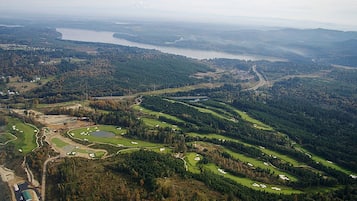 An aerial view of a golf course with multiple greens, a large body of water in the background, and surrounding forested areas.
