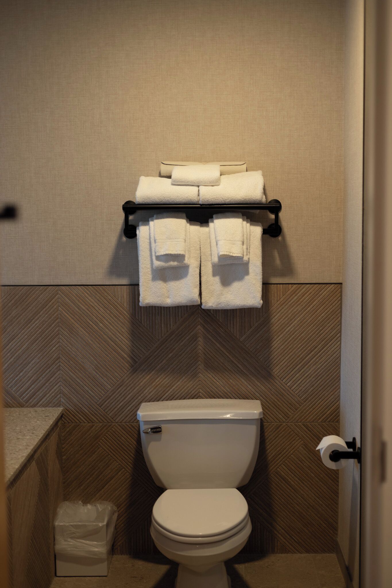 A bathroom with a toilet, a towel rack with folded towels, and a trash bin.