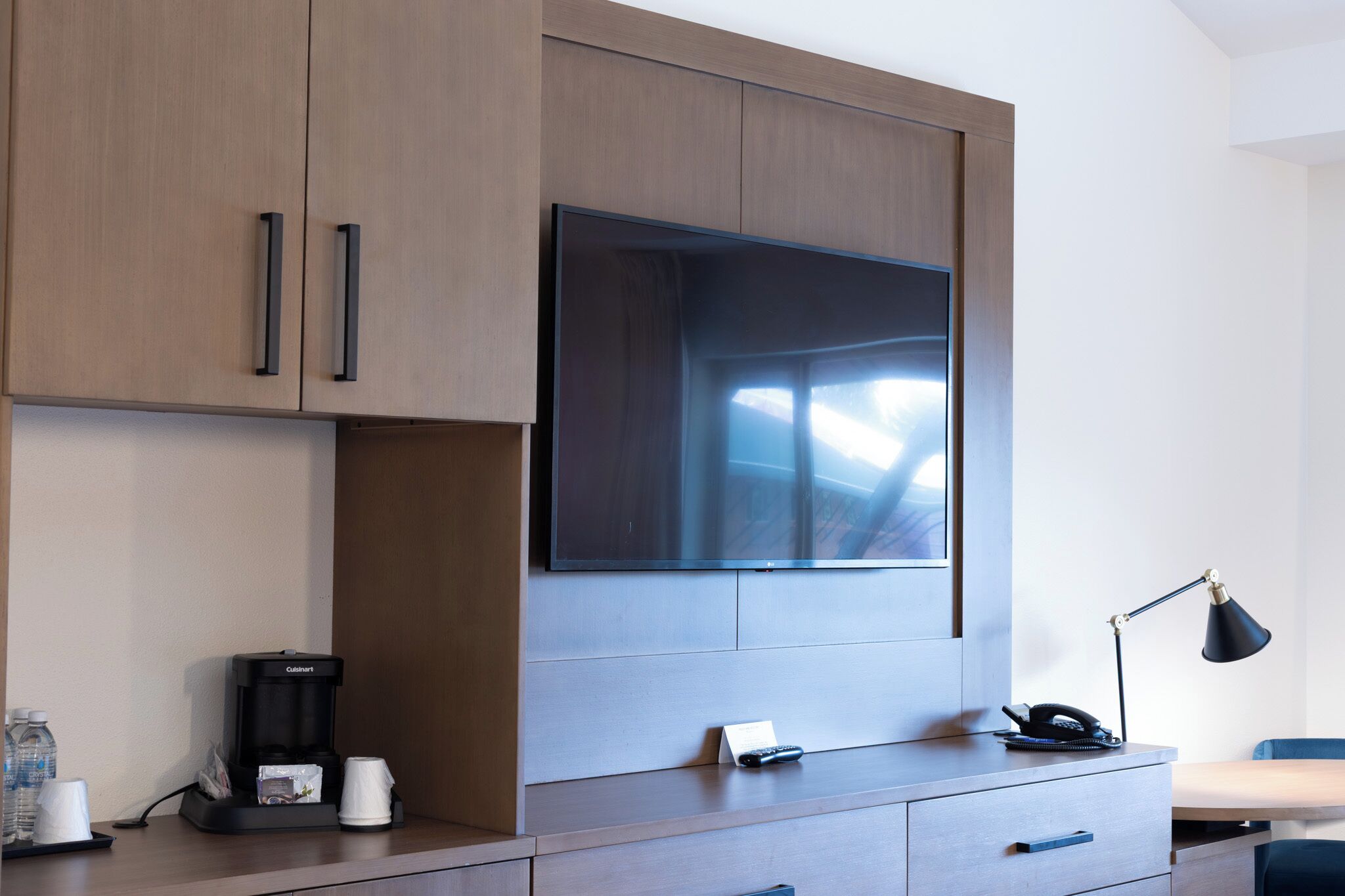 A modern hotel room with a large flat-screen TV mounted on a wooden cabinet, a built-in coffee maker, a desk with a lamp, and a chair.