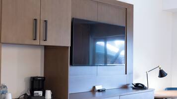 A modern hotel room with a large flat-screen TV mounted on a wooden cabinet, a built-in coffee maker, a desk with a lamp, and a chair.