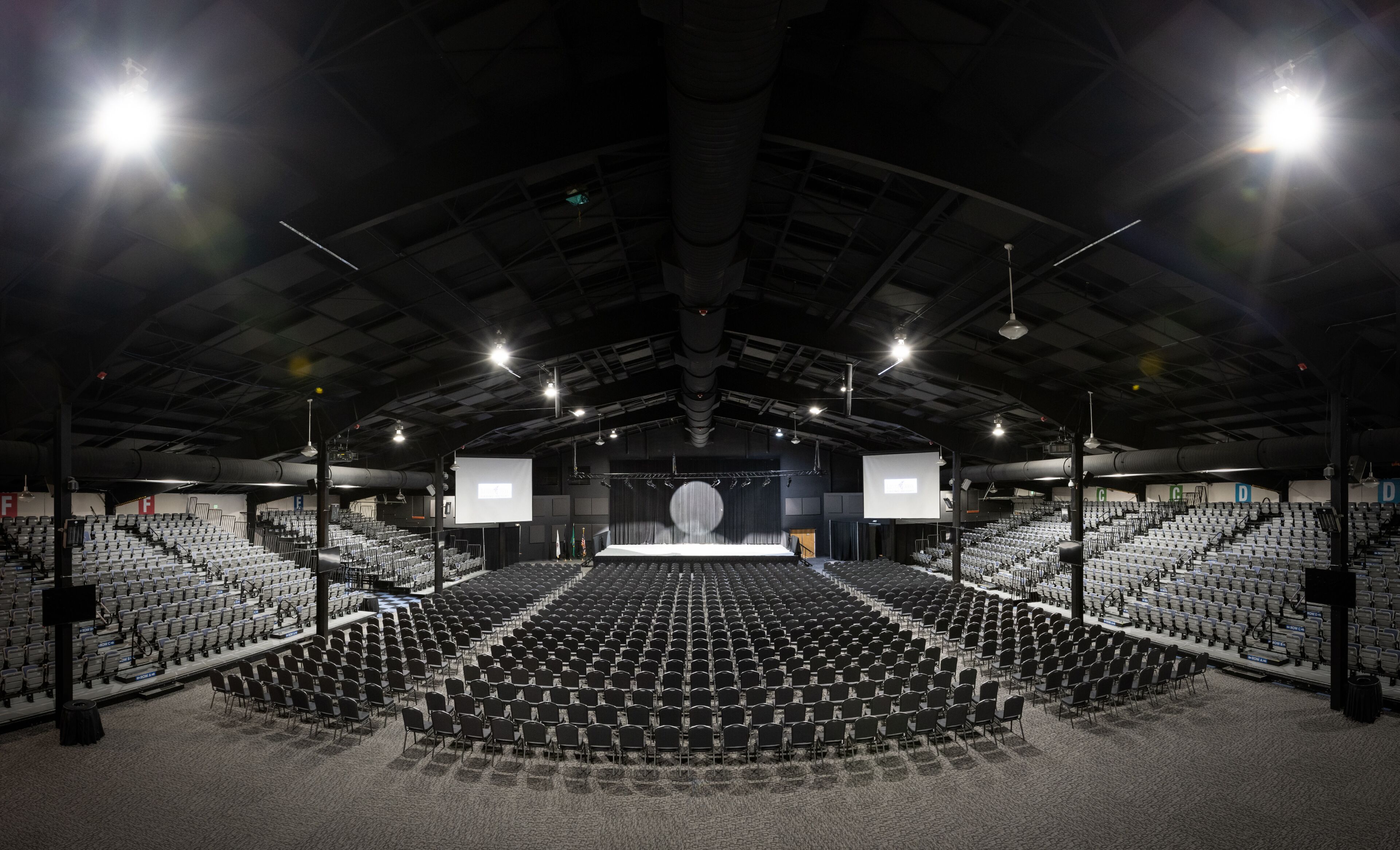 Een grote, lege aula met rijen stoelen, een podium en meerdere schermen.