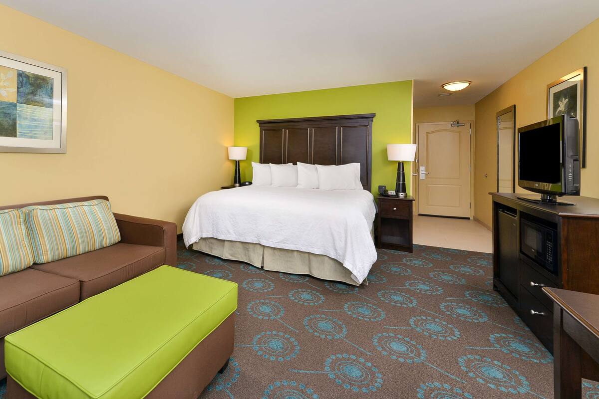 A hotel room with a bed, a green ottoman, a brown sofa, a television, and a door.