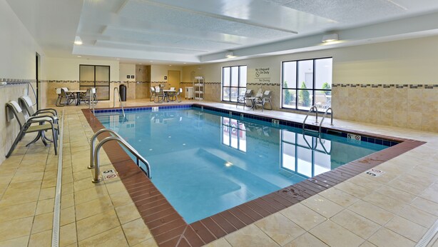 An indoor swimming pool with clear water, tiled walls, and a tiled floor. There are lounge chairs and a table with chairs around the pool.