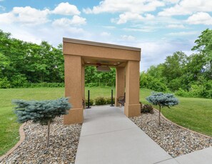 A concrete pathway leading to a beige stone archway with a bench under it, surrounded by gravel and small trees. - Hampton Inn Iowa City/University Area (Iowa City)