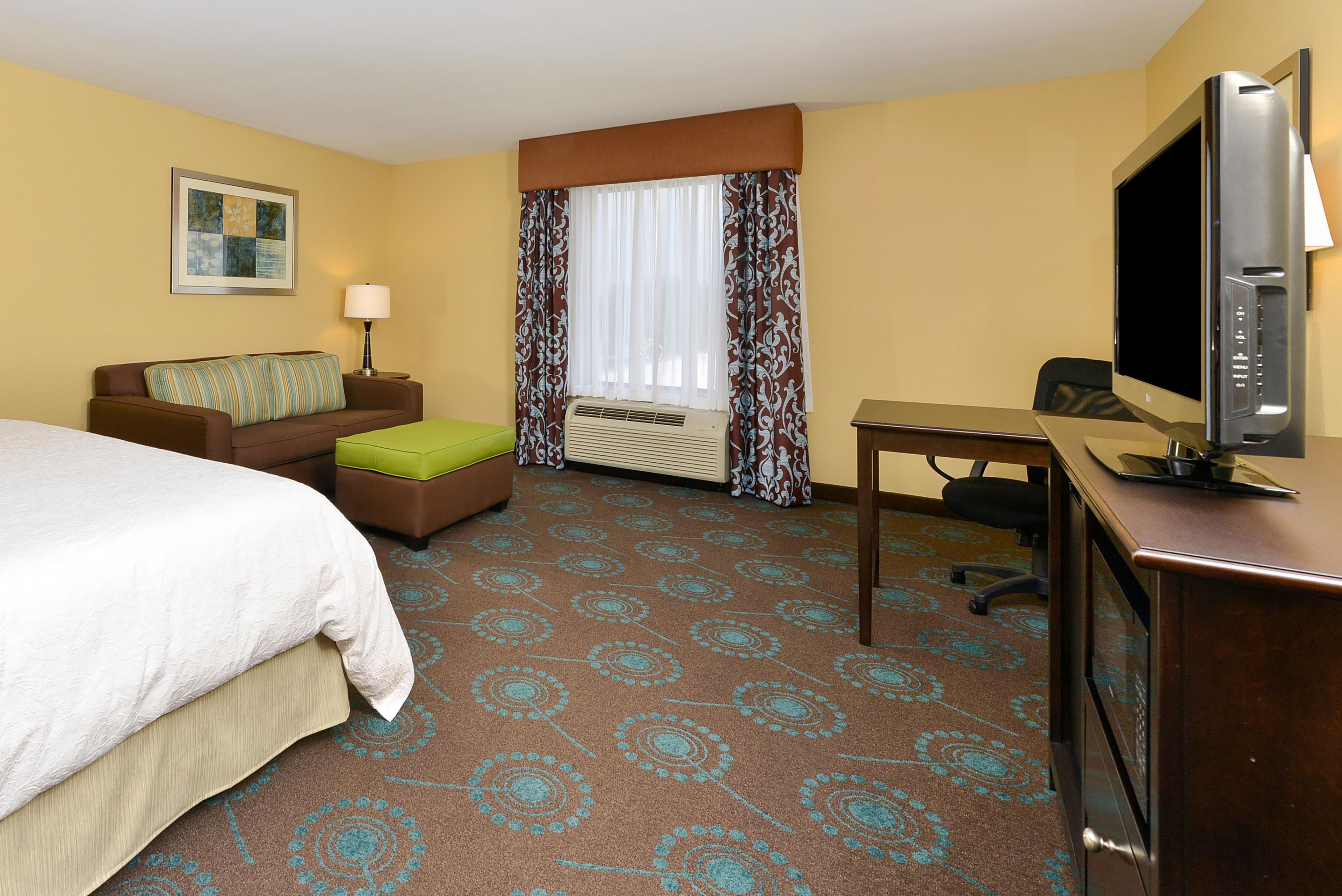 A hotel room with a bed, a desk, a television, a window with curtains, a wall-mounted lamp, and a framed picture.