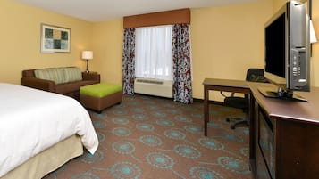 A hotel room with a bed, a desk, a television, a window with curtains, a wall-mounted lamp, and a framed picture.