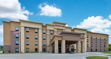 Un hotel Hampton Inn con un diseño moderno, que presenta una entrada central con pilares de piedra, varios pisos y un techo plano.