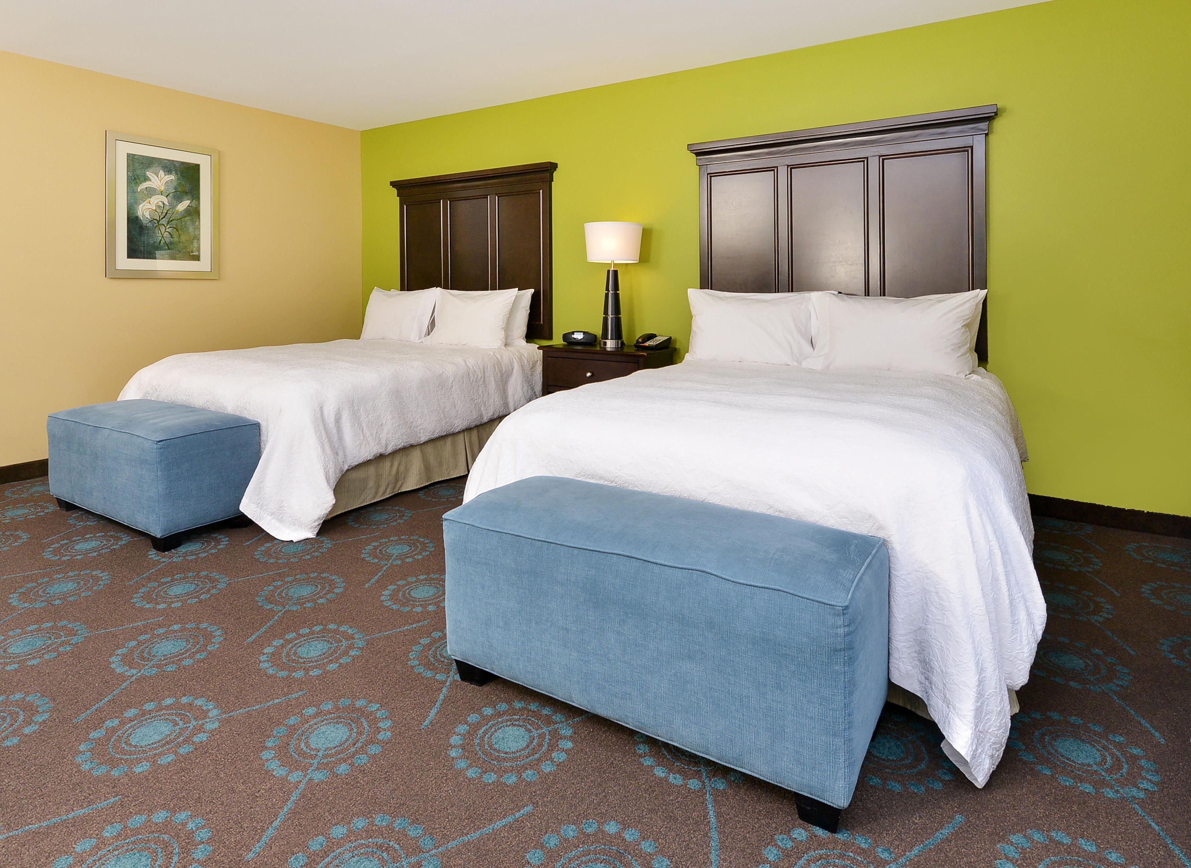 Quarto de hotel com duas camas, uma mesinha de cabeceira, uma luminária e um quadro na parede.