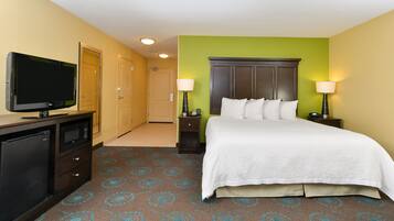 A hotel room with a bed, a TV, a desk with a lamp, and a door.