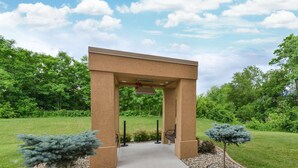 A covered walkway with a beige structure, surrounded by gravel and small trees, leading to a grassy area.