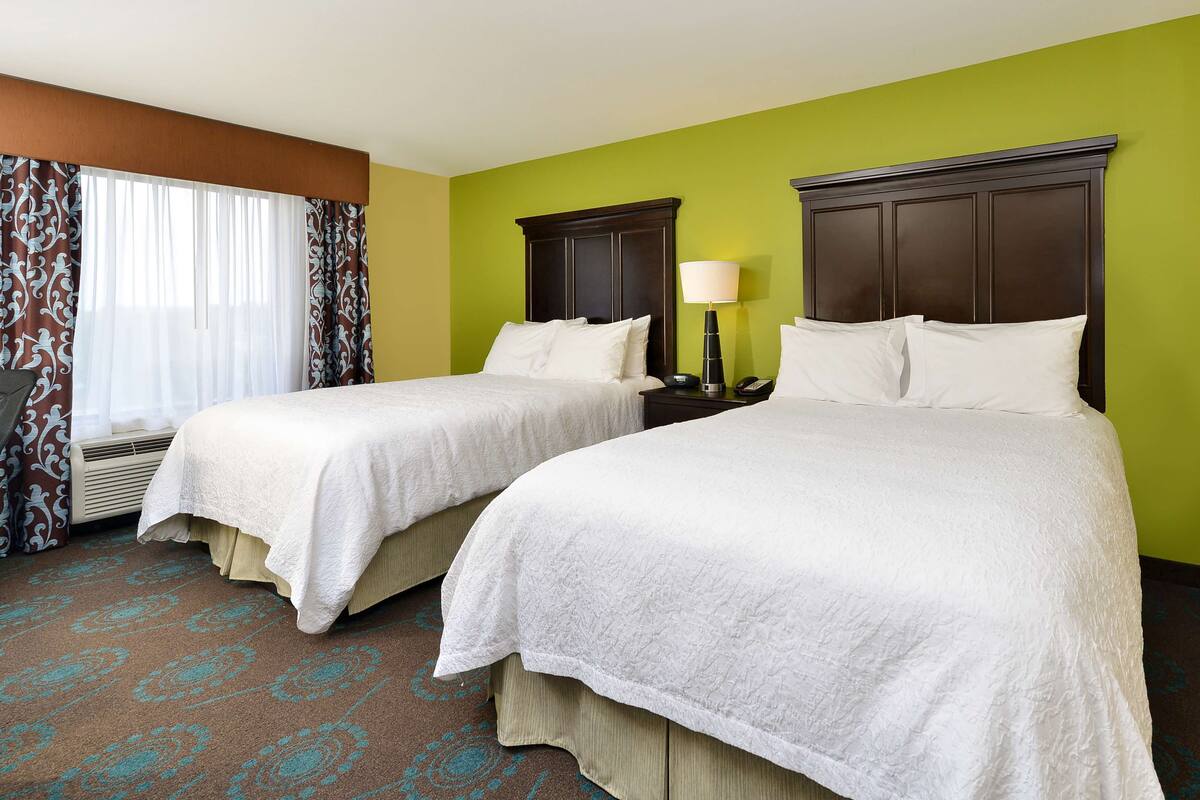 A hotel room with two beds, a window with curtains, a bedside table with a lamp, and a carpet with a geometric pattern.