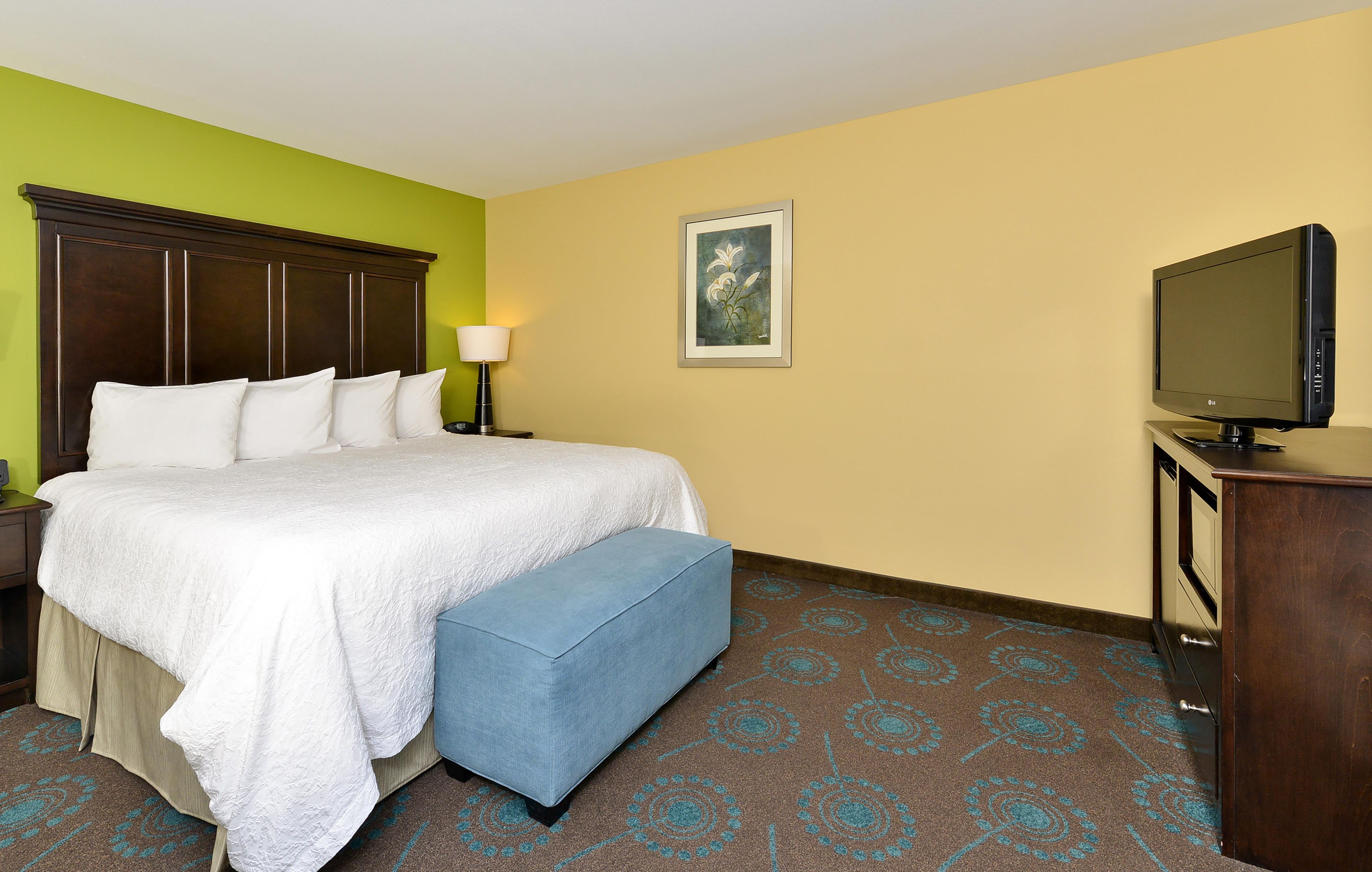 A hotel room with a bed, a bench, a television, and a painting on the wall.