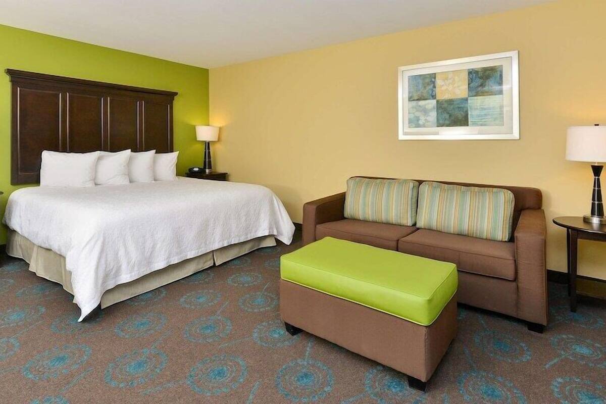 Una habitación de hotel con una cama, un sofá, un puf verde, un cuadro en la pared y dos lámparas de noche.