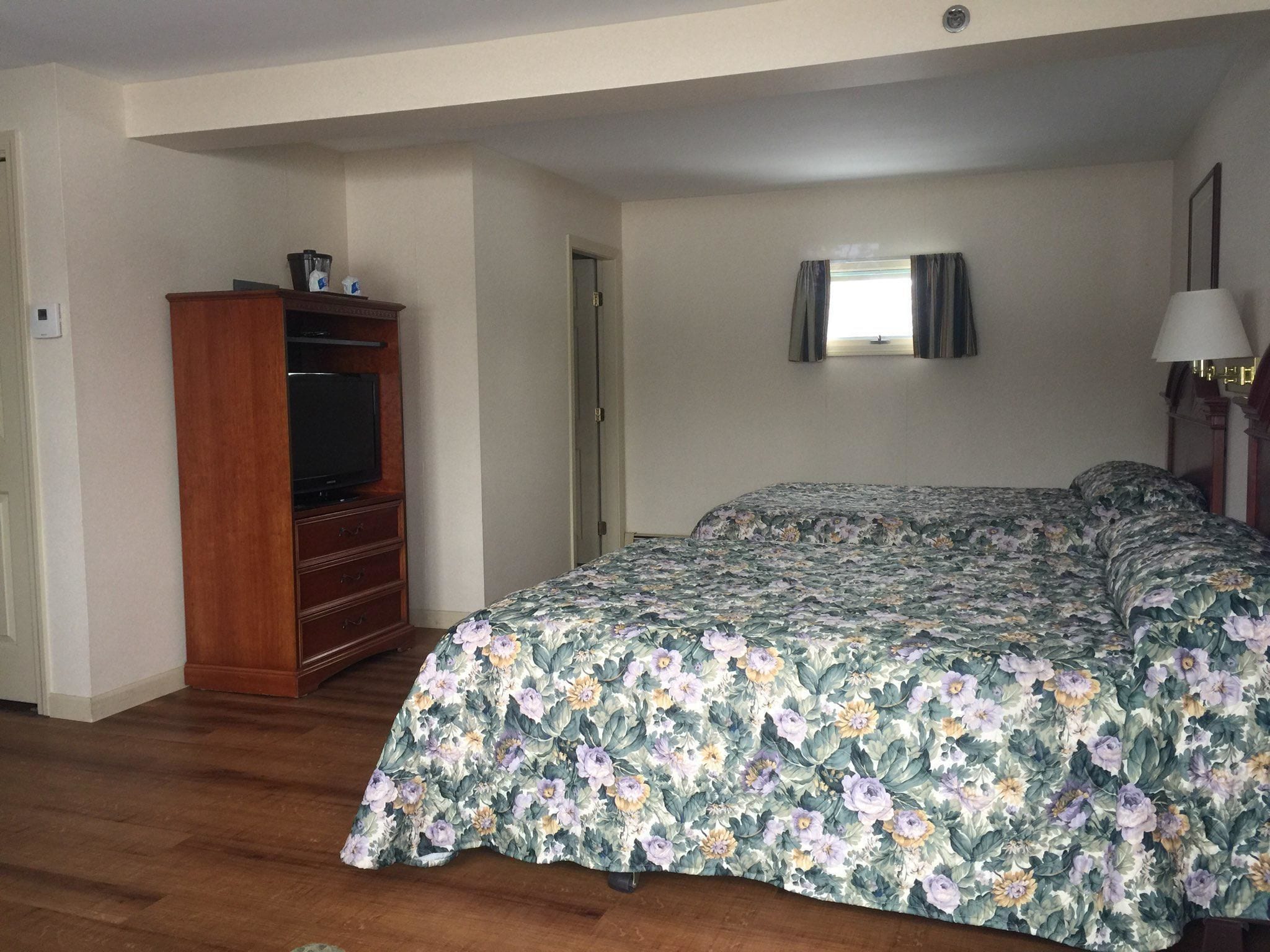 A bedroom with a wooden dresser, a television, a bed with floral bedding, a bedside lamp, and a window with curtains.