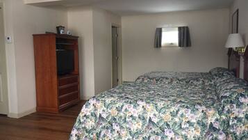 A bedroom with a wooden dresser, a television, a bed with floral bedding, a bedside lamp, and a window with curtains.