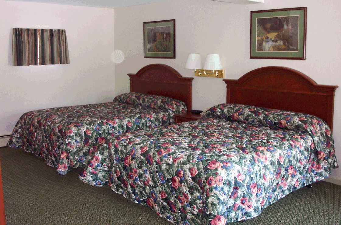 Quarto de hotel com duas camas de solteiro, uma mesa de cabeceira com abajures e duas fotografias emolduradas na parede.