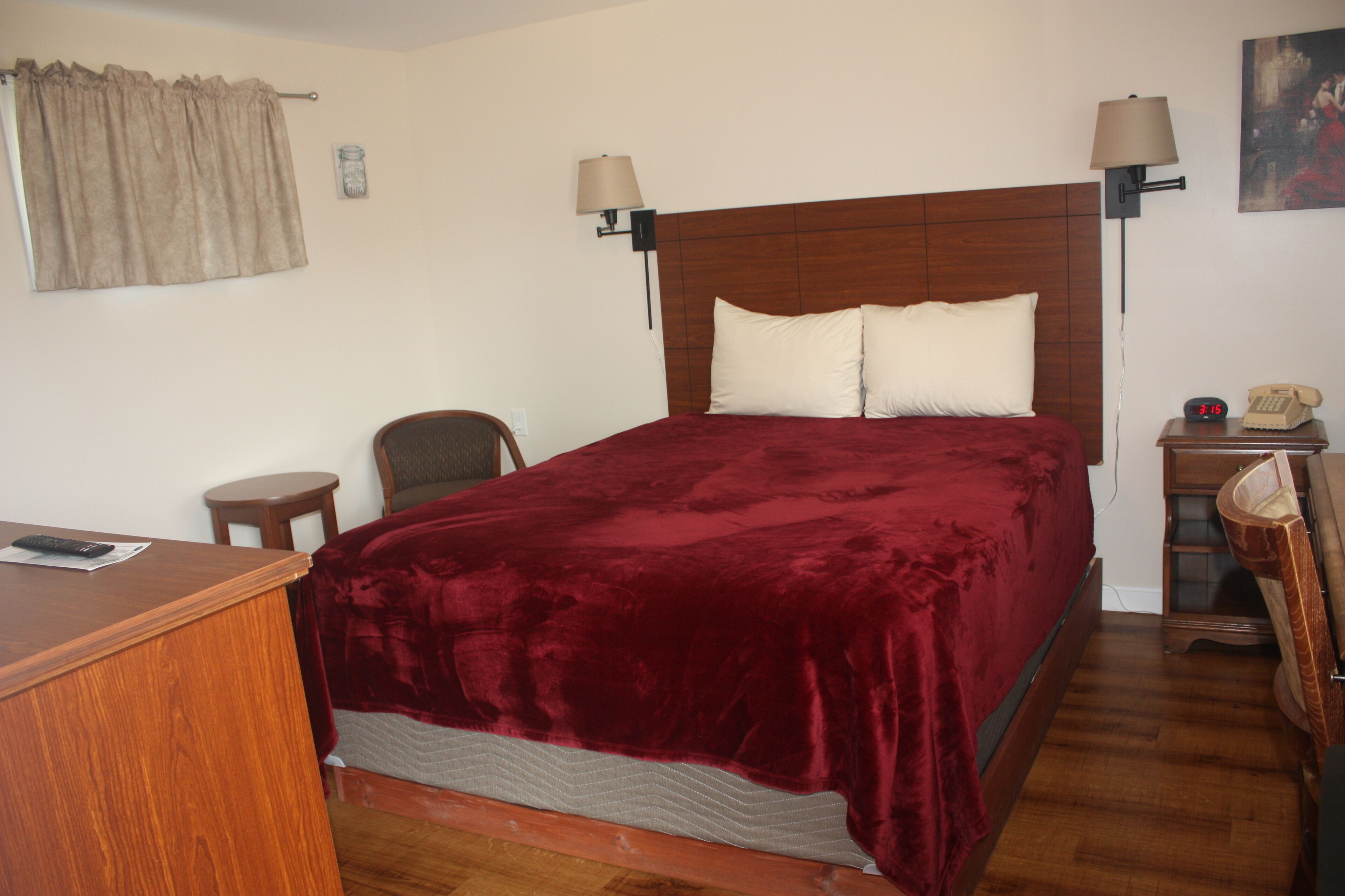 A hotel room with a wooden headboard, a red velvet bedspread, two bedside lamps, a small wooden nightstand with a telephone, and a chair.