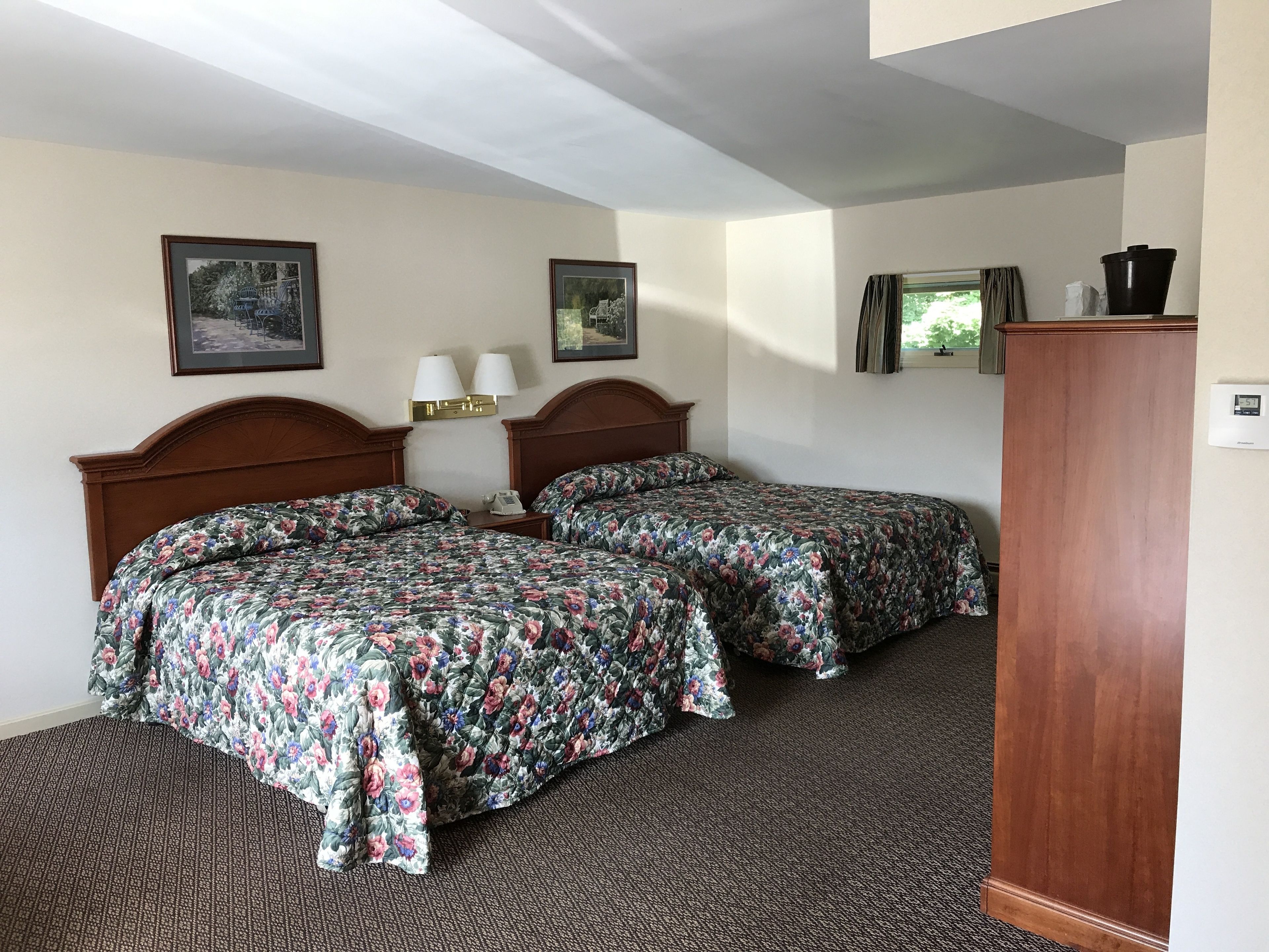 A hotel room with two beds, a wooden headboard, a nightstand with a lamp, and a thermostat on the wall.
