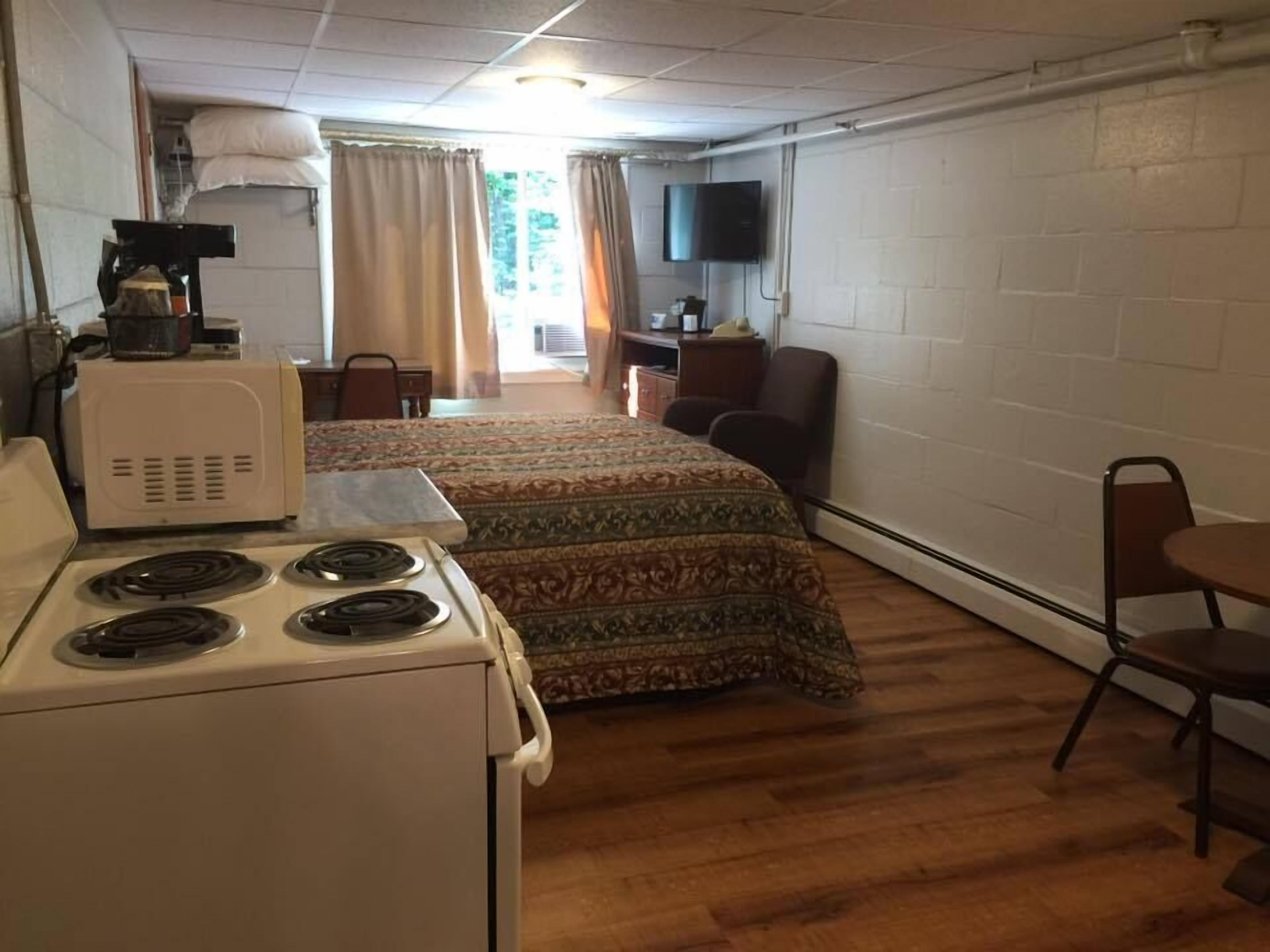 A compact room with a kitchenette, a bed with patterned bedding, a television mounted on the wall, and a window with curtains.