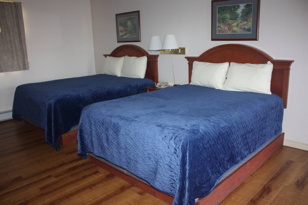 A hotel room with two beds, wooden flooring, and a nightstand with a lamp.