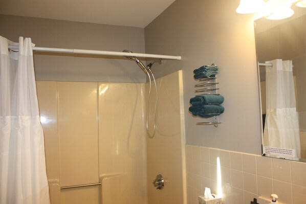 A bathroom with a shower, a towel rack with towels, a toilet paper holder, and a mirror. - The Towne Motel (Skowhegan)