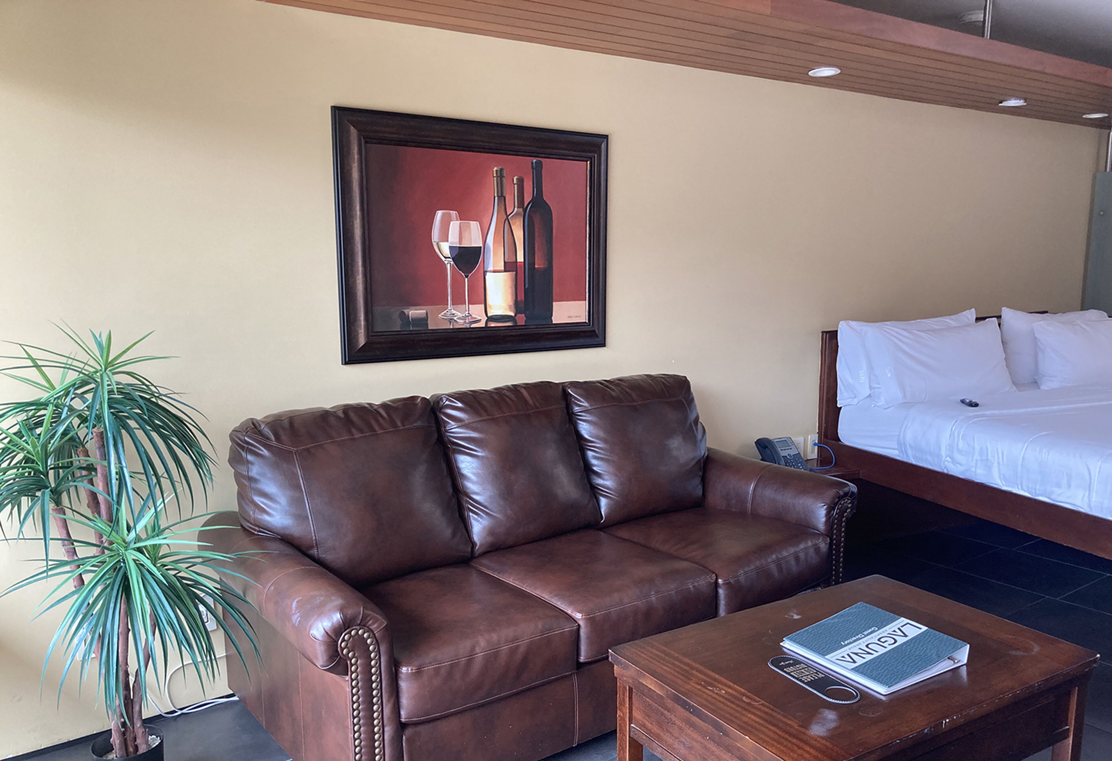 A brown leather sofa, a wooden coffee table with a book, a potted plant, and a bed with white linens.