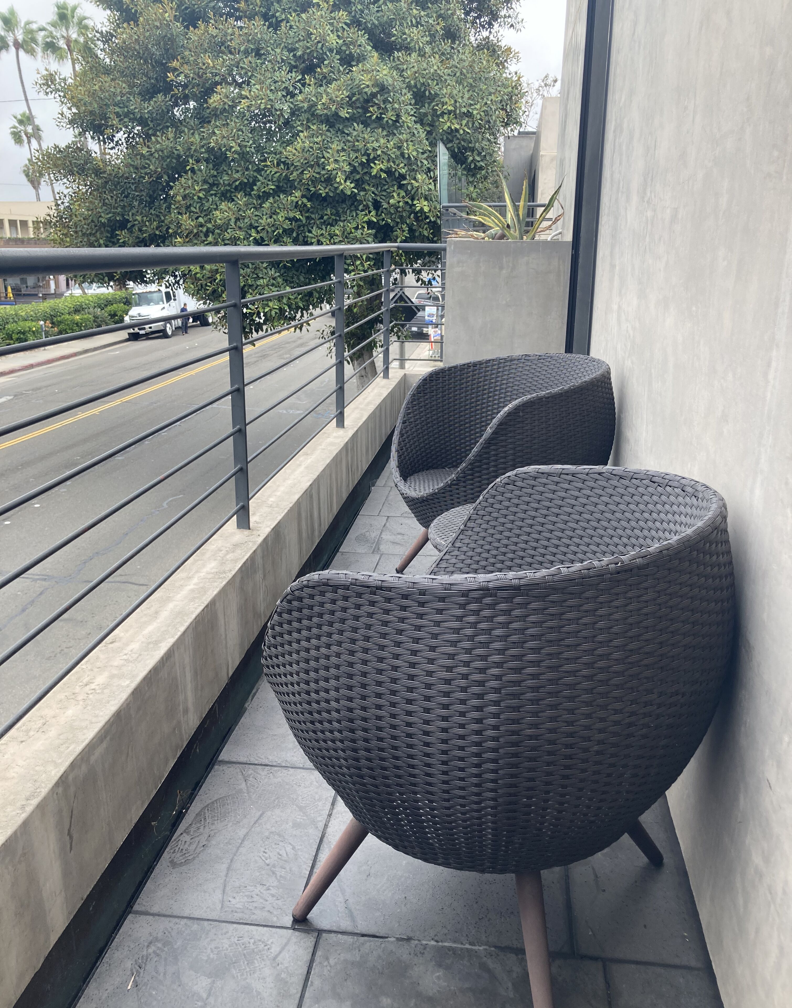 Un balcón con dos sillones de mimbre, piso de concreto, barandilla de metal y vista a una calle con un camión blanco.