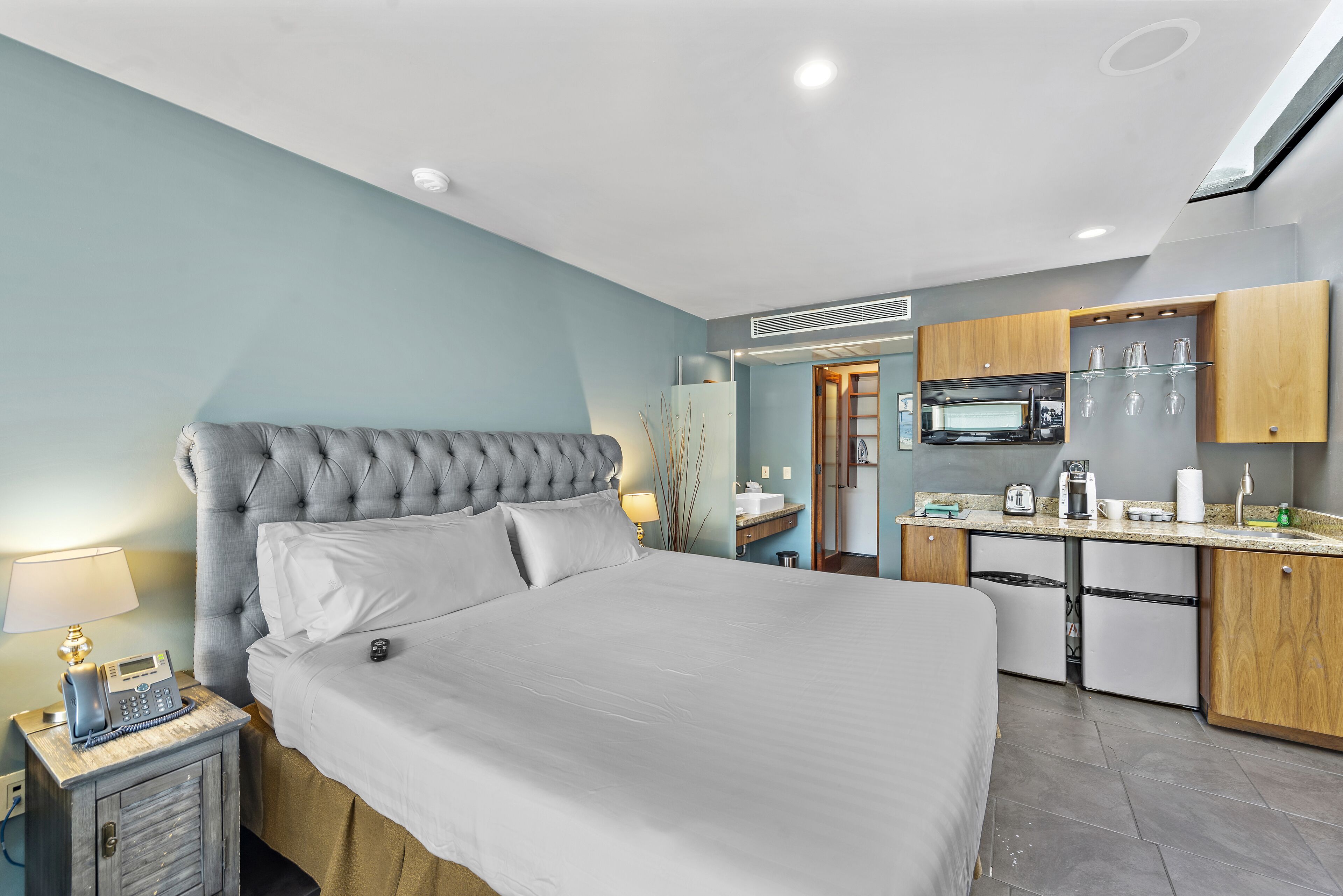 Quarto de hotel moderno com uma cama grande, telefone ao lado da cama, uma pequena mesa com luminária, e uma cozinha compacta com micro-ondas, cafeteira e armários.