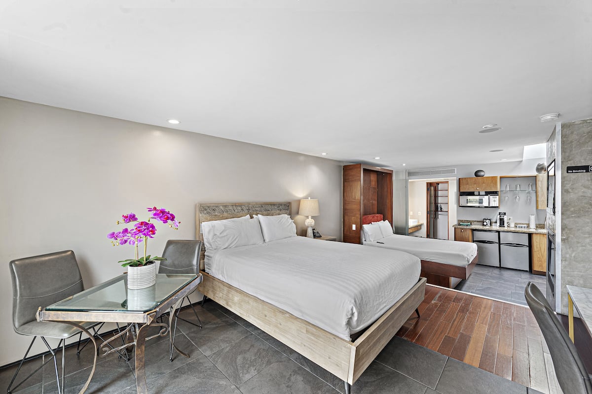 A modern hotel room with a large bed, a small desk with a chair, a glass table with a vase of flowers, and a kitchenette in the background.