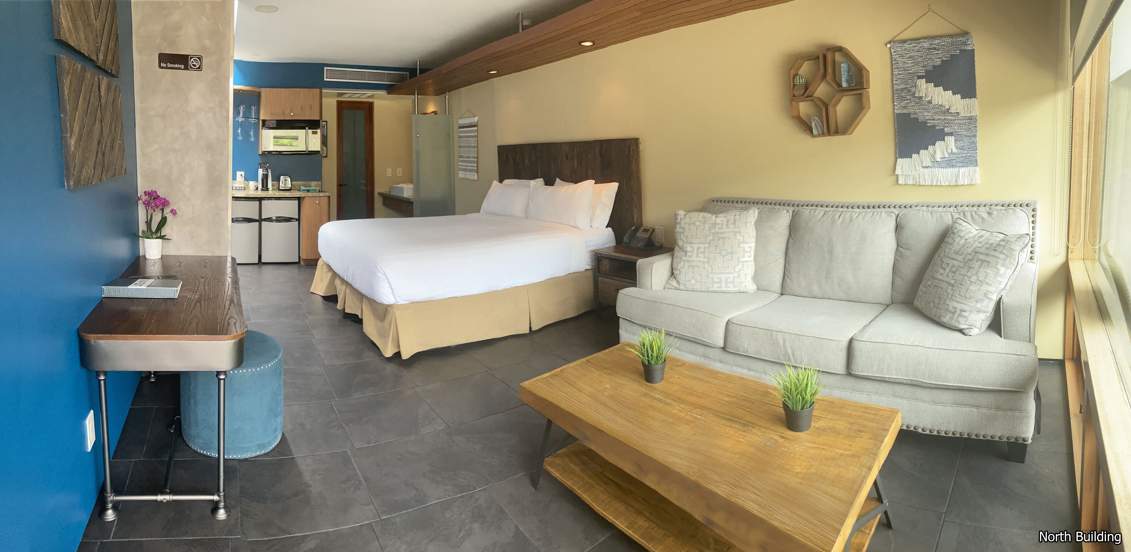 A hotel room with a bed, a sofa, a coffee table, a small table with a plant, and a kitchenette in the background.
