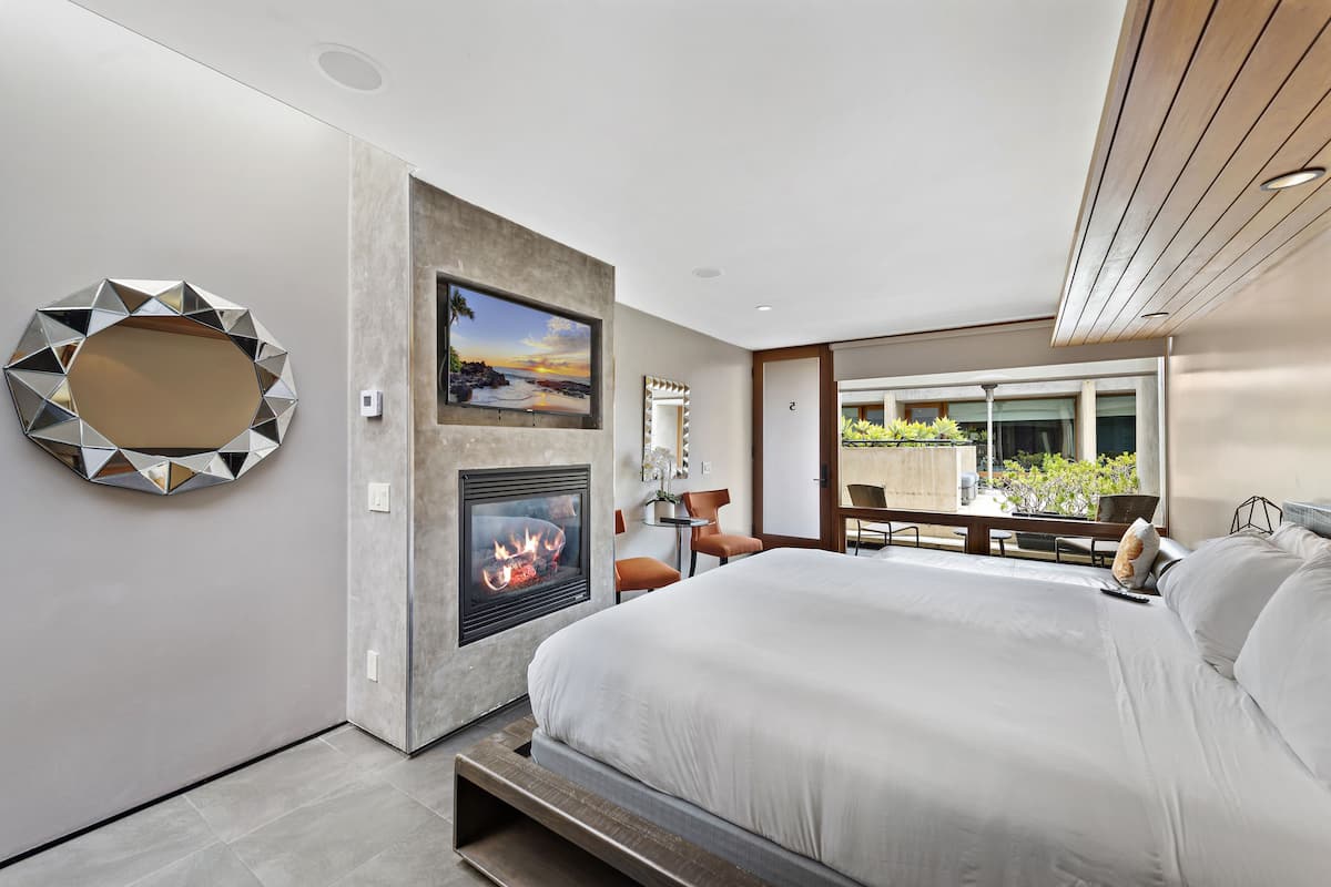 A modern bedroom with a large bed, a fireplace, a TV, and a balcony with plants.