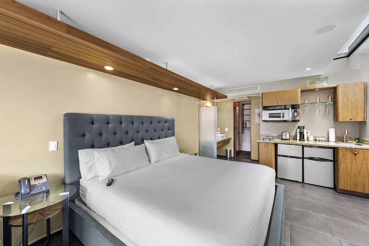 A modern hotel room with a large bed, a small bedside table with a phone, a kitchenette with a microwave, and a glass cabinet with glasses.