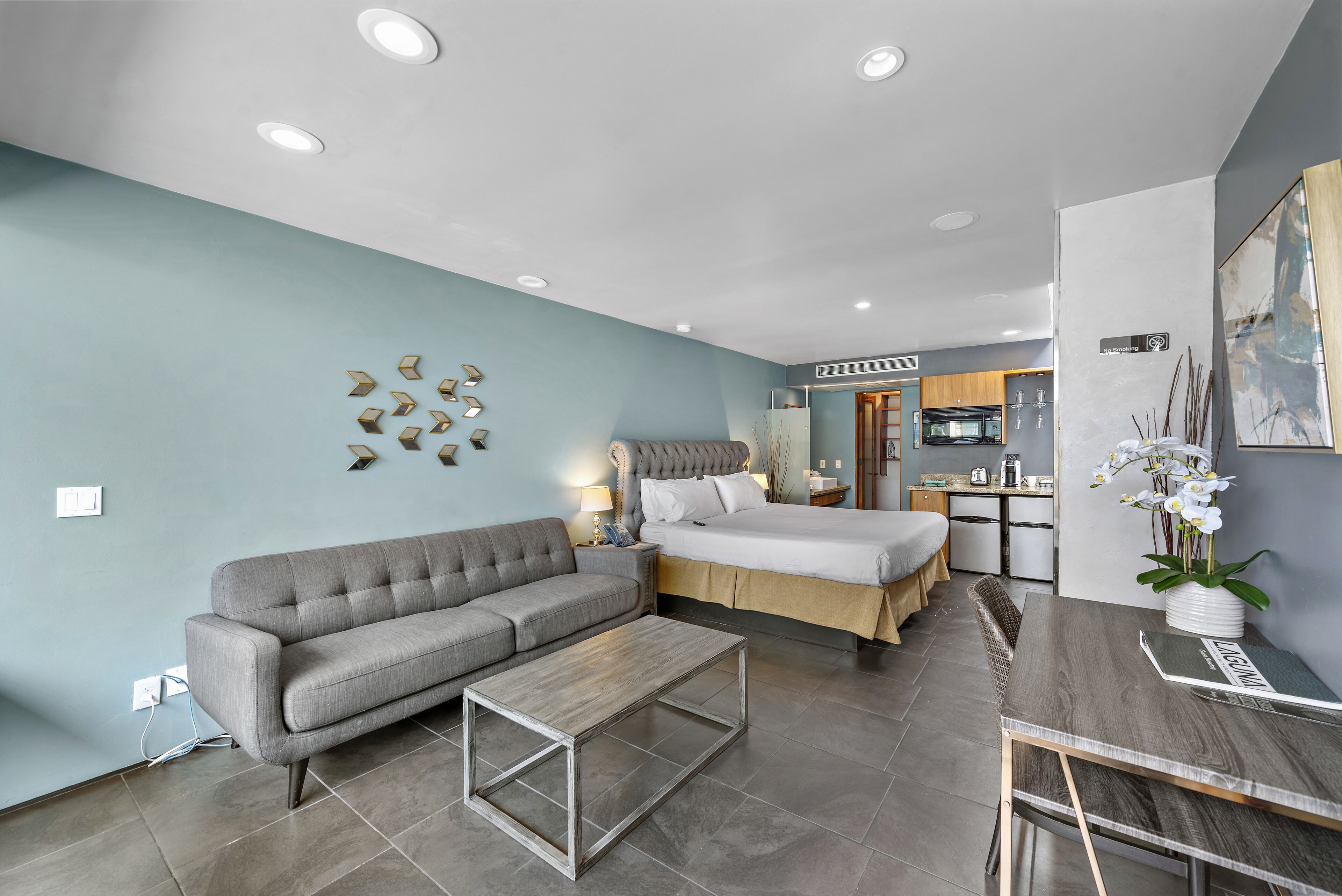 Une chambre d’hôtel moderne avec un canapé gris, une table basse en bois, un lit avec une tête de lit grise, un petit coin cuisine et une table à manger.